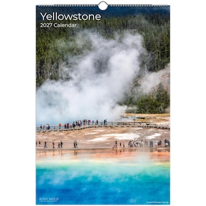 May include: A vertical wall calendar titled Yellowstone 2027 Calendar featuring a photograph of the Grand Prismatic Spring. The image displays a vibrant blue pool, orange mineral deposits, a wooden boardwalk with people, and rising steam.