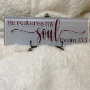 May include: A rectangular glass plaque with the words "He restores my soul" in a burgundy script font, with "Psalm 23:3" below. The plaque is displayed on a small black metal stand.
