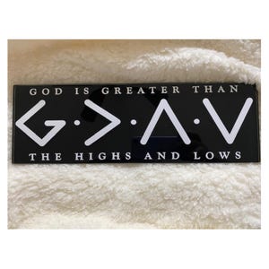 May include: Black rectangular sign with white text that reads "GOD IS GREATER THAN G > A V THE HIGHS AND LOWS". The sign is on a white, fluffy surface.