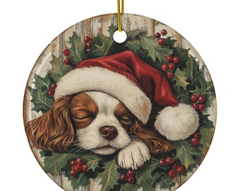 May include: A round Christmas ornament featuring a sleeping Cavalier King Charles Spaniel wearing a Santa hat. The dog rests on a bed of holly and red berries, set against a rustic wood-grain background. A gold string is attached for hanging.