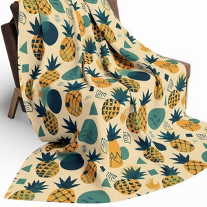 May include: A beige throw blanket draped over a chair, featuring a repeating pattern of yellow and green pineapples, blue shapes, and geometric designs. The blanket is made of a soft material and is suitable for home decor.