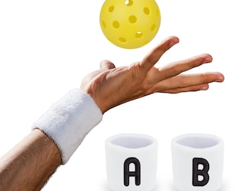 May include: A yellow pickleball with multiple holes is suspended above a hand wearing a white wristband. Two white wristbands with the letters "A" and "B" are below the hand. The wristbands are made of a soft, absorbent material.