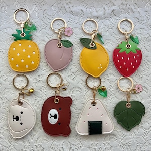Peut inclure: Un ensemble de dix porte-clés colorés avec différents motifs, dont un ananas, une pêche, un citron, une fraise, un ours, un onigiri et une feuille. Chaque porte-clés a un anneau en or et un petit charme.