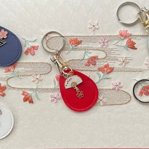 May include: Five keychains with different colored leather teardrop-shaped charms. Each charm has a gold metal fan charm with a pink flower charm attached. The charms are on a silver key ring.
