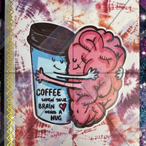May include: A notebook with a tie-dye background featuring a graphic of a blue coffee cup hugging a pink brain. The text reads: COFFEE WHEN YOUR BRAIN NEEDS A HUG, with a small red heart. A gold zigzag pattern runs along the left edge of the cover.
