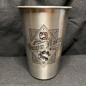 Puede incluir: Un vaso de metal plateado cepillado con la parte superior acampanada. El vaso presenta un emblema decorativo con las palabras "Dungeon Master" y dos ilustraciones de dragones. El diseño está grabado en color marrón oscuro.