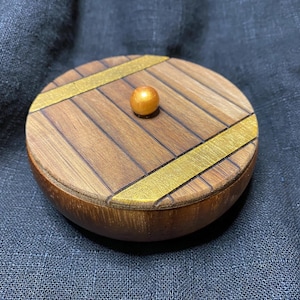 May include: A round wooden box with a lid. The lid is made of wooden planks with gold-colored bands and a small gold sphere on top. The box is brown and sits on a dark blue surface.