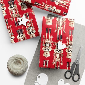 Op de afbeelding: Rood cadeaupapier met een herhalend patroon van skeletnotenkrakers, vastgebonden met touw en een stervormig label. Een kluwen wit garen, een schaar en blanco labels zijn ook aanwezig.