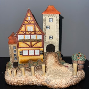 May include: A miniature ceramic model of a German-style house with a tower, a cobblestone path, and a small flower pot with flowers. The house is painted in shades of brown, yellow, and white.