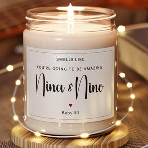 May include: A white candle with a label that reads "Smells like you're going to be amazing Nina & Nino Baby Jill". The candle is lit and sitting on a wooden coaster.