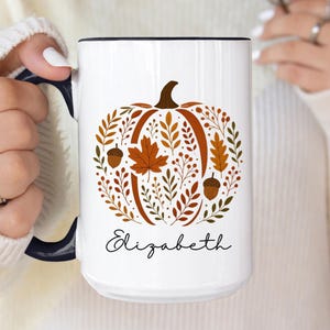 May include: A white ceramic mug with a black handle and interior rim. The front features a graphic of a pumpkin composed of autumn leaves, branches, and acorns in shades of orange and brown. The name Elizabeth is printed in black script below.
