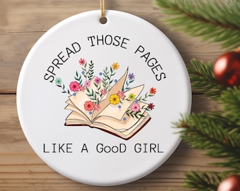 May include: White ceramic ornament with the text "SPREAD THOSE PAGES LIKE A GOOD GIRL" in black. The ornament features an open book illustration with colorful flowers and greenery. A gold string is attached for hanging.