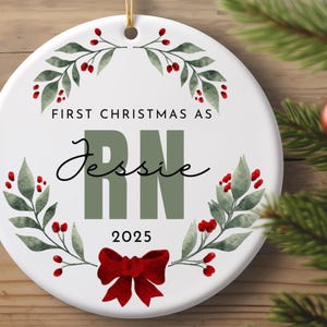 May include: A white ceramic Christmas ornament with the text "FIRST CHRISTMAS AS Jessie RN 2025". The ornament features a green and red floral design, a red bow, and a gold string for hanging. Festive holiday decor.