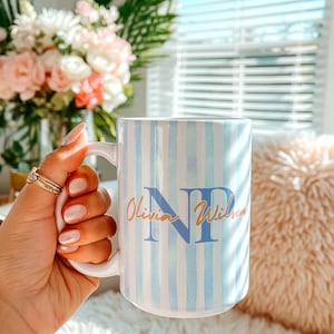 May include: White ceramic mug with blue and white vertical stripes. The mug features the initials "NP" in blue and the name "Olivia Wilson" in gold script. The mug is held in a hand with multiple rings.