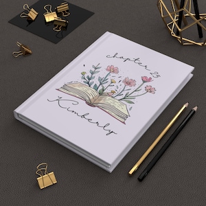 May include: White hardcover journal with an open book illustration featuring pink flowers and greenery. The cover includes the text "chapter 25" and "Kimberly" in script. Gold and black pencils and gold binder clips are nearby.