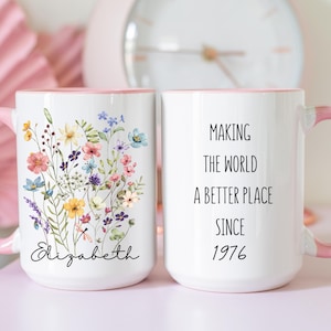 May include: White ceramic mug with a pink handle and rim. One side features a floral design with the name "Elizabeth". The other side reads "Making the world a better place since 1976".