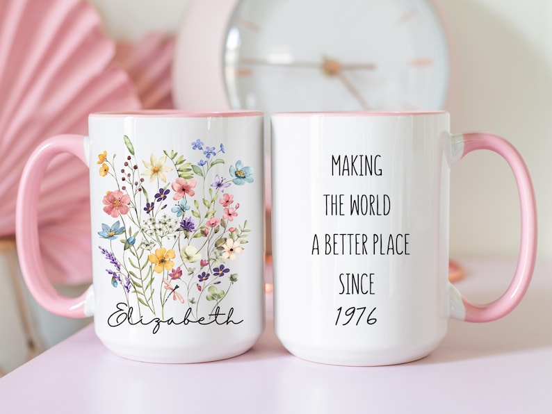 May include: White ceramic mug with a pink handle and rim. One side features a floral design with the name "Elizabeth". The other side reads "Making the world a better place since 1976".
