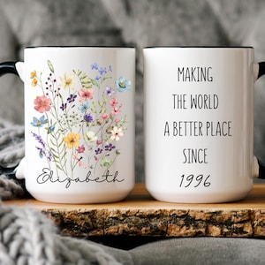May include: White ceramic mug with a black handle and rim. One side features a floral design with colorful flowers and the name "Elizabeth". The other side reads "MAKING THE WORLD A BETTER PLACE SINCE 1996".