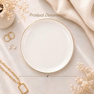May include: A circular white ceramic trinket dish featuring a thin gold rim. The dish has a diameter of 4 inches as indicated by the text and measurement line. The surface is smooth and glossy, designed for holding small items like jewellery.