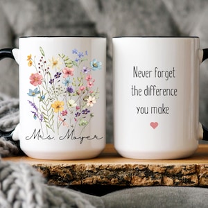 May include: White ceramic mug with a black handle and rim. One side features a floral design with colorful wildflowers and the name "Mrs. Mayer". The other side has the text "Never forget the difference you make" with a pink heart.