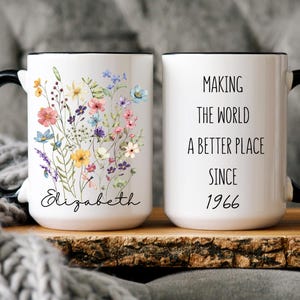 May include: A white ceramic mug with a black handle and rim. One side features a colorful watercolor wildflower bouquet above the name Elizabeth in black script. The other side displays the black text Making the world a better place since 1966.