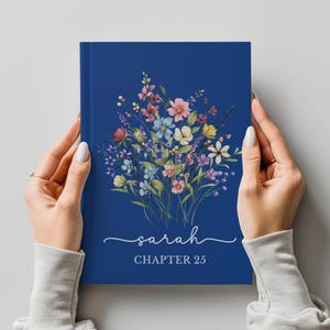 May include: A navy blue journal with a floral bouquet design. The bouquet features pink, yellow, blue, and white flowers. The name "Sarah" is written in cursive, with "CHAPTER 25" below it.
