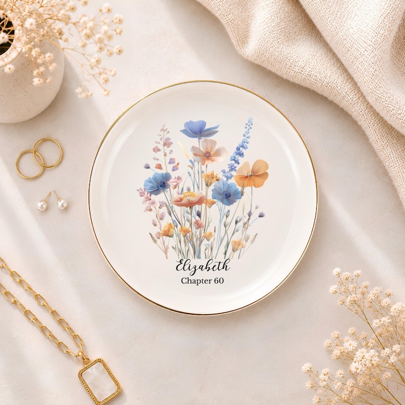 May include: A round white ceramic trinket dish with a gold-coloured rim. The centre features a watercolour-style illustration of blue, orange, and pink wildflowers. Below the flowers, the text reads Elizabeth and Chapter 60 in black script and print.