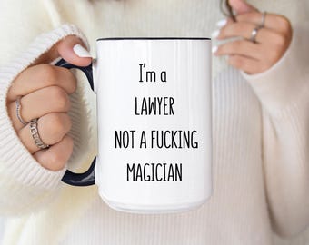 May include: White ceramic mug with a black handle and rim. The mug features the text "I'm a LAWYER NOT A FUCKING MAGICIAN" in black, bold font. A humorous gift for a lawyer.