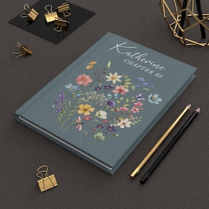 May include: A teal journal with the name "Katherine" and "Chapter 25" in white script. The cover features a floral bouquet in shades of pink, yellow, blue, and purple. Gold and black pencils and binder clips are also visible.