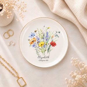 May include: A round white ceramic trinket dish with a gold-toned rim, featuring a colourful watercolour illustration of wildflowers in shades of blue, yellow, orange, and red. The text "Elizabeth" and "Chapter 60" is printed in black script and serif font.