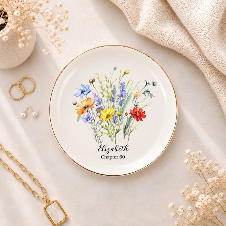 May include: A round white ceramic trinket dish with a gold-toned rim, featuring a colourful watercolour illustration of wildflowers in shades of blue, yellow, orange, and red. The text "Elizabeth" and "Chapter 60" is printed in black script and serif font.