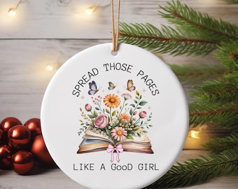 May include: White ceramic ornament with a watercolor illustration of an open book, flowers, and butterflies. The text "SPREAD THOSE PAGES LIKE A GOOD GIRL" is printed around the image. A brown string is attached for hanging.