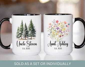 May include: Two white ceramic mugs with black handles and black rims. The mug on the left has a forest scene with the text "Uncle Steven Est. 2025". The mug on the right has a bouquet of colorful flowers with the text "Aunt Ashley Est. 2025".