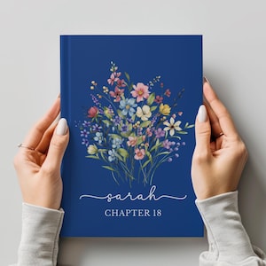 May include: A navy blue journal with a watercolor floral design. The cover features a bouquet of pink, blue, yellow, and purple flowers. The name "Sarah" is written in cursive, with "CHAPTER 18" below it.