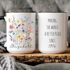 May include: White ceramic mug with a black handle and rim. One side features a floral design with various colors and the name "Elizabeth". The other side reads "MAKING THE WORLD A BETTER PLACE SINCE 1976".