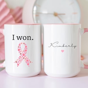 May include: Two white ceramic mugs with pink handles and rims. One mug has the text "I won." and a pink floral ribbon design. The other mug has the name "Kimberly" and a small pink heart.