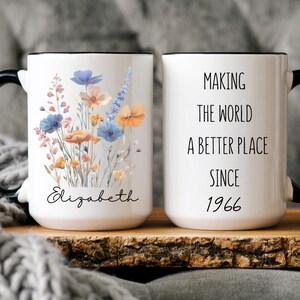 May include: A white ceramic mug with a black handle and rim. One side features a colourful wildflower bouquet illustration above the name Elizabeth in black script. The other side displays the black text Making the world a better place since 1966.