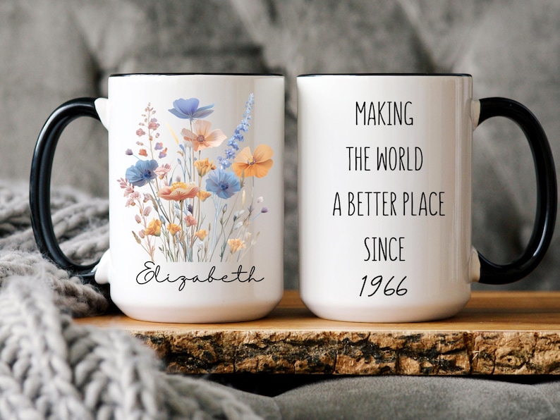 May include: A white ceramic mug with a black handle and rim. One side features a colourful wildflower bouquet illustration above the name Elizabeth in black script. The other side displays the black text Making the world a better place since 1966.