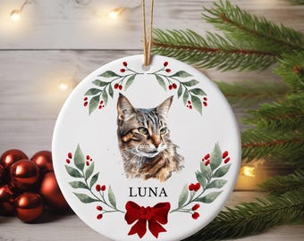 May include: A round, white ceramic Christmas ornament featuring a watercolor portrait of a tabby cat. The cat's name, "LUNA," is printed below the image. The ornament is decorated with a green and red berry wreath and a red bow.