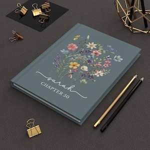 May include: A teal journal with a floral design and the name "sarah" and "CHAPTER 50" in white script. Gold and black pencils and gold binder clips are on a dark gray surface.