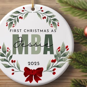 May include: A white ceramic Christmas ornament with the text "FIRST CHRISTMAS AS MBA Jessie 2025". The ornament is decorated with a watercolor wreath of green leaves and red berries, and a red bow.