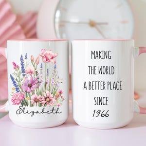 May include: Two white ceramic mugs with pink handles and rims. One mug features a colourful floral bouquet illustration above the name Elizabeth in black script. The other mug displays the black text Making the world a better place since 1966.