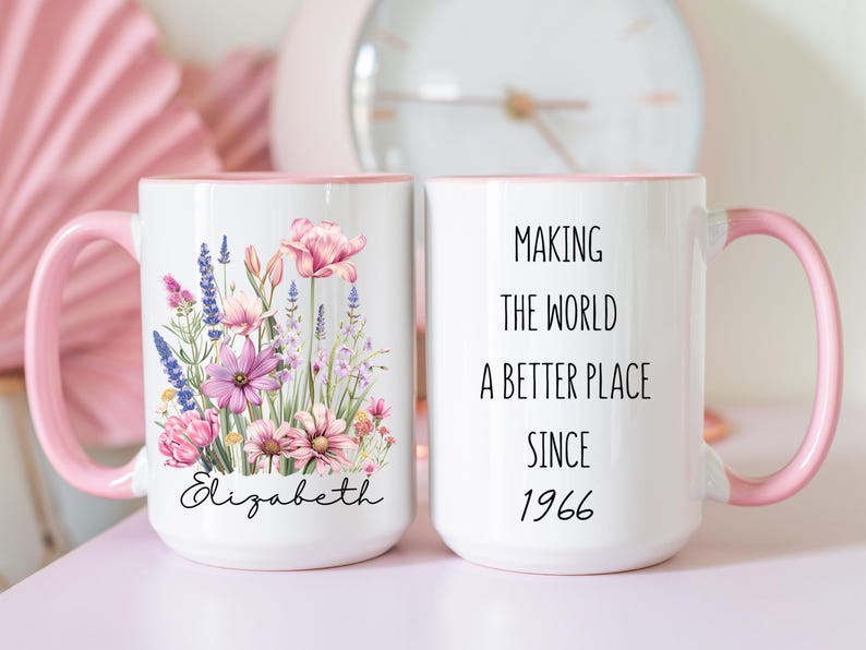 May include: Two white ceramic mugs with pink handles and rims. One mug features a colourful floral bouquet illustration above the name Elizabeth in black script. The other mug displays the black text Making the world a better place since 1966.