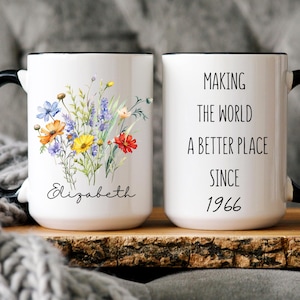 May include: A white ceramic mug with a black handle and rim. One side features a colourful wildflower bouquet and the name Elizabeth in script. The other side displays the text Making the world a better place since 1966 in a simple black font.