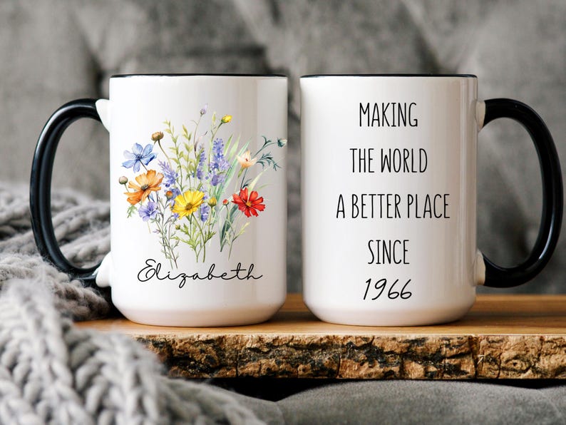 May include: A white ceramic mug with a black handle and rim. One side features a colourful wildflower bouquet and the name Elizabeth in script. The other side displays the text Making the world a better place since 1966 in a simple black font.