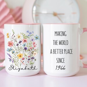 May include: Two white ceramic mugs with light pink handles and interior rims. One mug features a colourful wildflower bouquet illustration above the name Elizabeth in black script. The other mug displays the black text Making the world a better place since 1966.