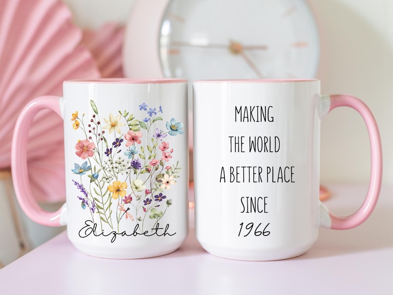 May include: Two white ceramic mugs with light pink handles and interior rims. One mug features a colourful wildflower bouquet illustration above the name Elizabeth in black script. The other mug displays the black text Making the world a better place since 1966.