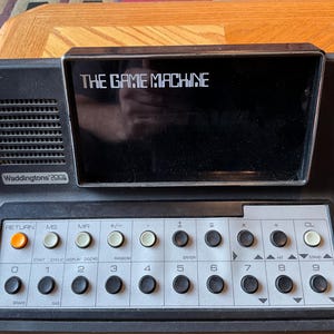 May include: A black and silver vintage electronic game console with a screen that reads "THE GAME MACHINE". The console has a keypad with buttons labeled "RETURN", "MS", "MR", "+/-", "-", "=", "x", "CL", "START", "CYCLE", "DISPLAY", "ZIGZAG", "RANDOM", "ENTER", "4", "5", "6", "7", "8", "9", "0", "1", "2", "3", and "STAND".