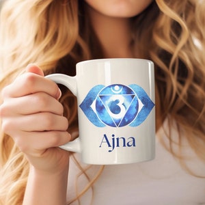 May include: White ceramic mug with a blue and white watercolor design of the third eye chakra, also known as the Ajna chakra. The design includes a stylized eye, a crescent moon, and the Om symbol. The word "Ajna" is written in blue below the design.