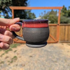 May include: A ceramic mug with a rounded body and a small handle. The lower portion of the mug features a matte, dark charcoal gray finish, while the upper rim is glazed in a vibrant, glossy red that transitions into a deep blue at the edge.